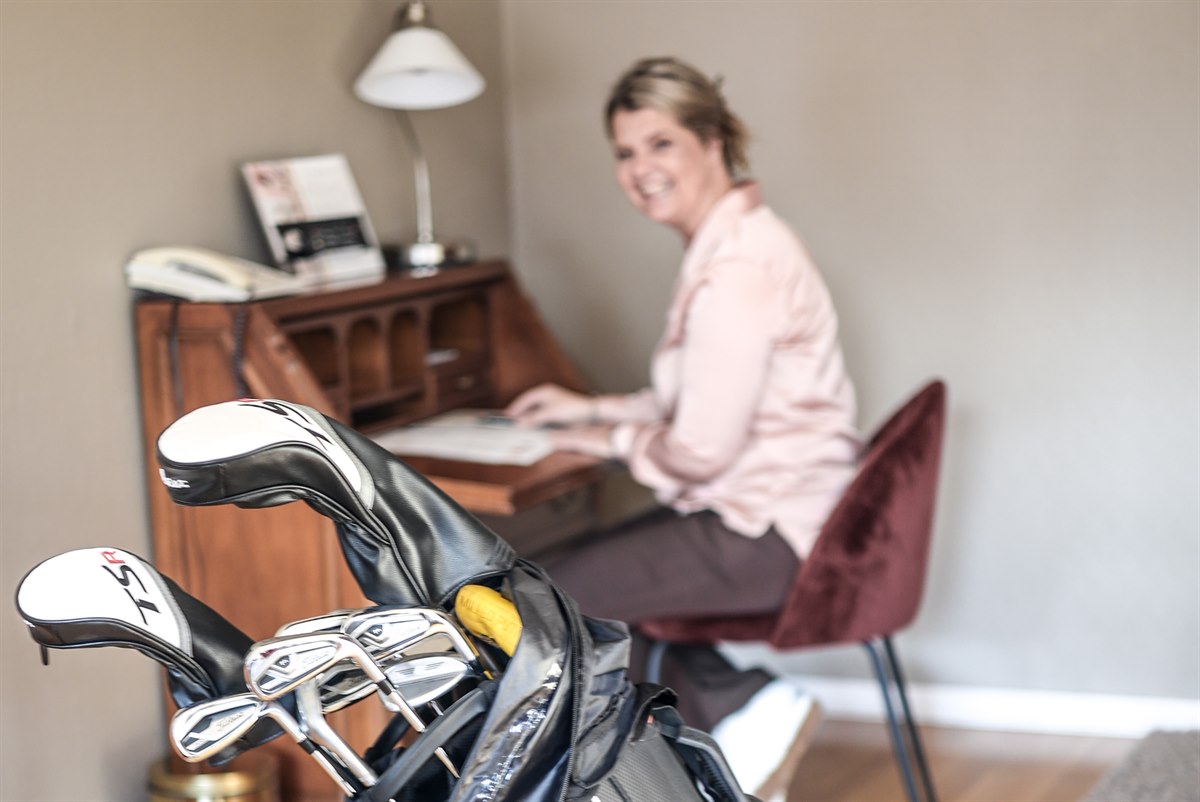 A golf bag and a woman at a desk in a hotel room.
